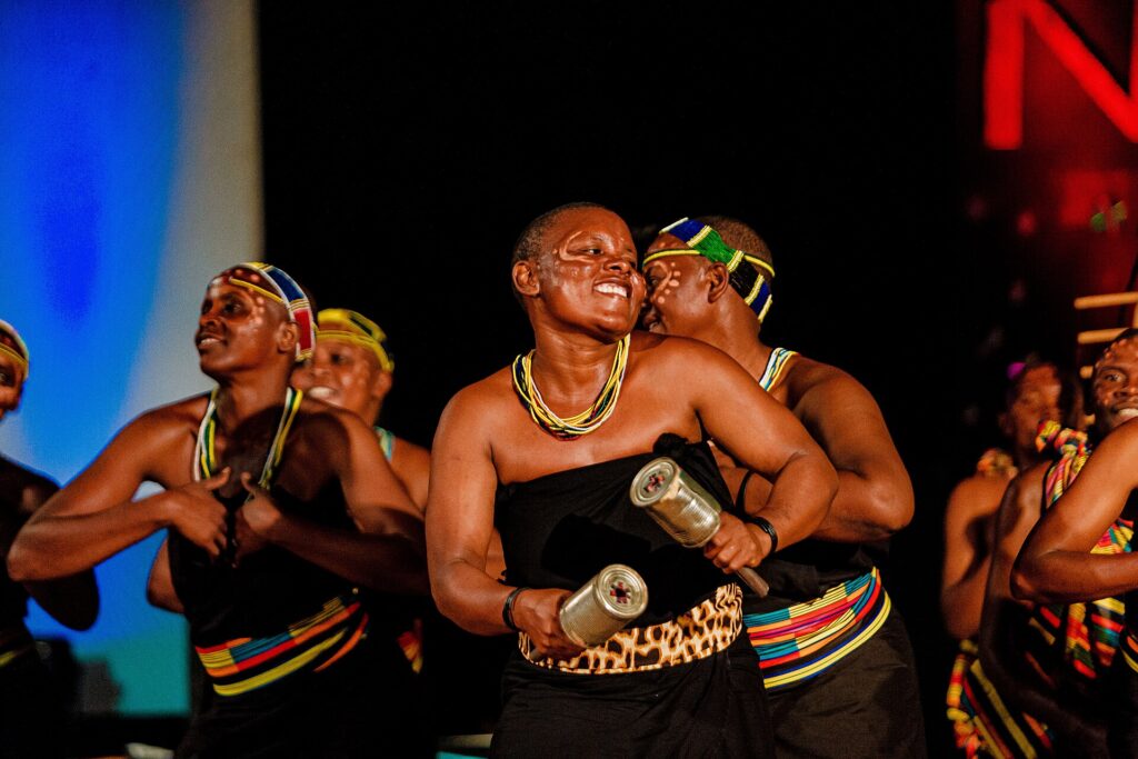 Gogo dancers and musicians performing on stage in Tanzania, one holding a pair of shakers